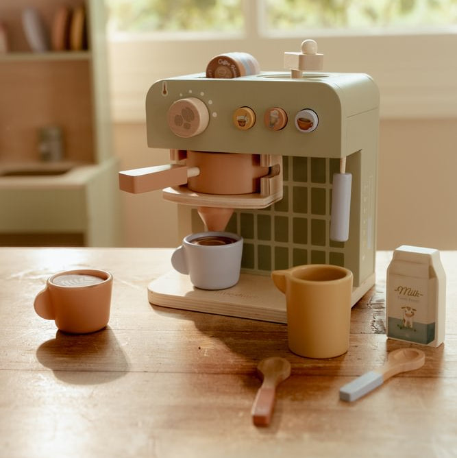 Grüne Little Dutch Kaffeemaschine mit hellblauer Tasse unter dem Auslauf, orangefarbener Tasse links und Milchkanne, Milchkarton und Löffeln auf dem Tisch.
