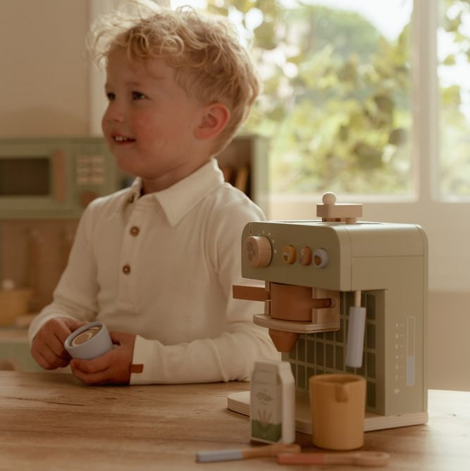 Kind hält eine hellblaue Spieltasse neben der grünen Little Dutch Kaffeemaschine mit gelber Milchkanne auf einem Holztisch.
