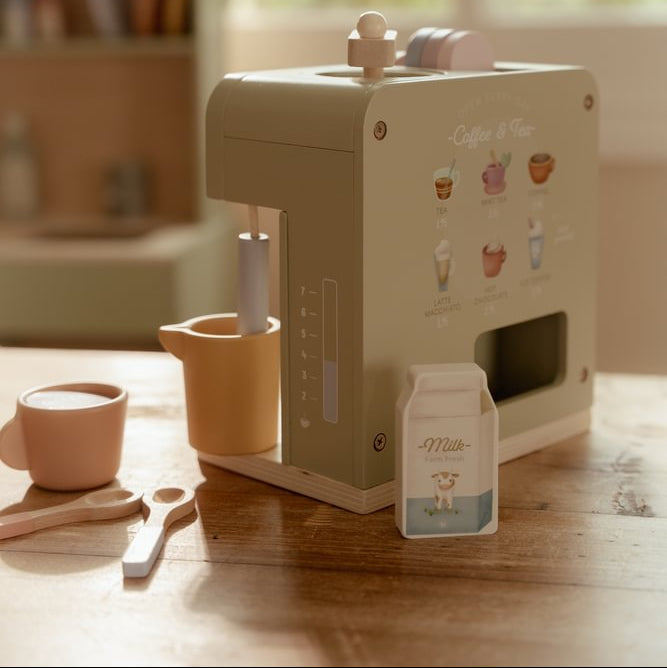 Schräge Rückansicht der grünen Little Dutch Kaffeemaschine mit Getränkemotiven, seitlicher Milchkanne und Milchkarton auf dem Holztisch.
